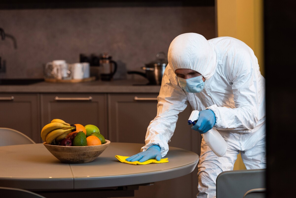Selective focus of man in hazmat suit and medical mask cleaning table in kitchen