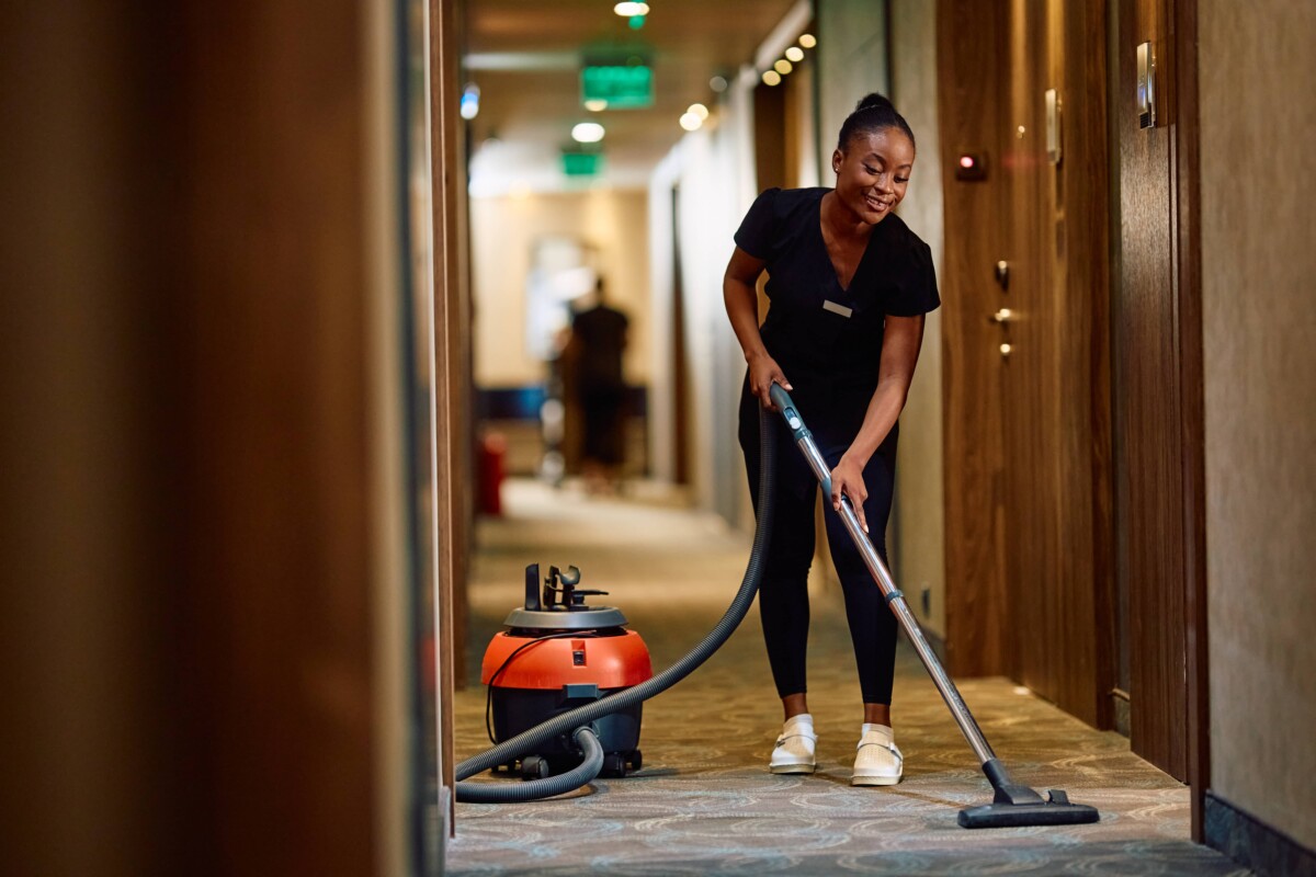 Young black maid vacuuming hallway while working in a hotel.